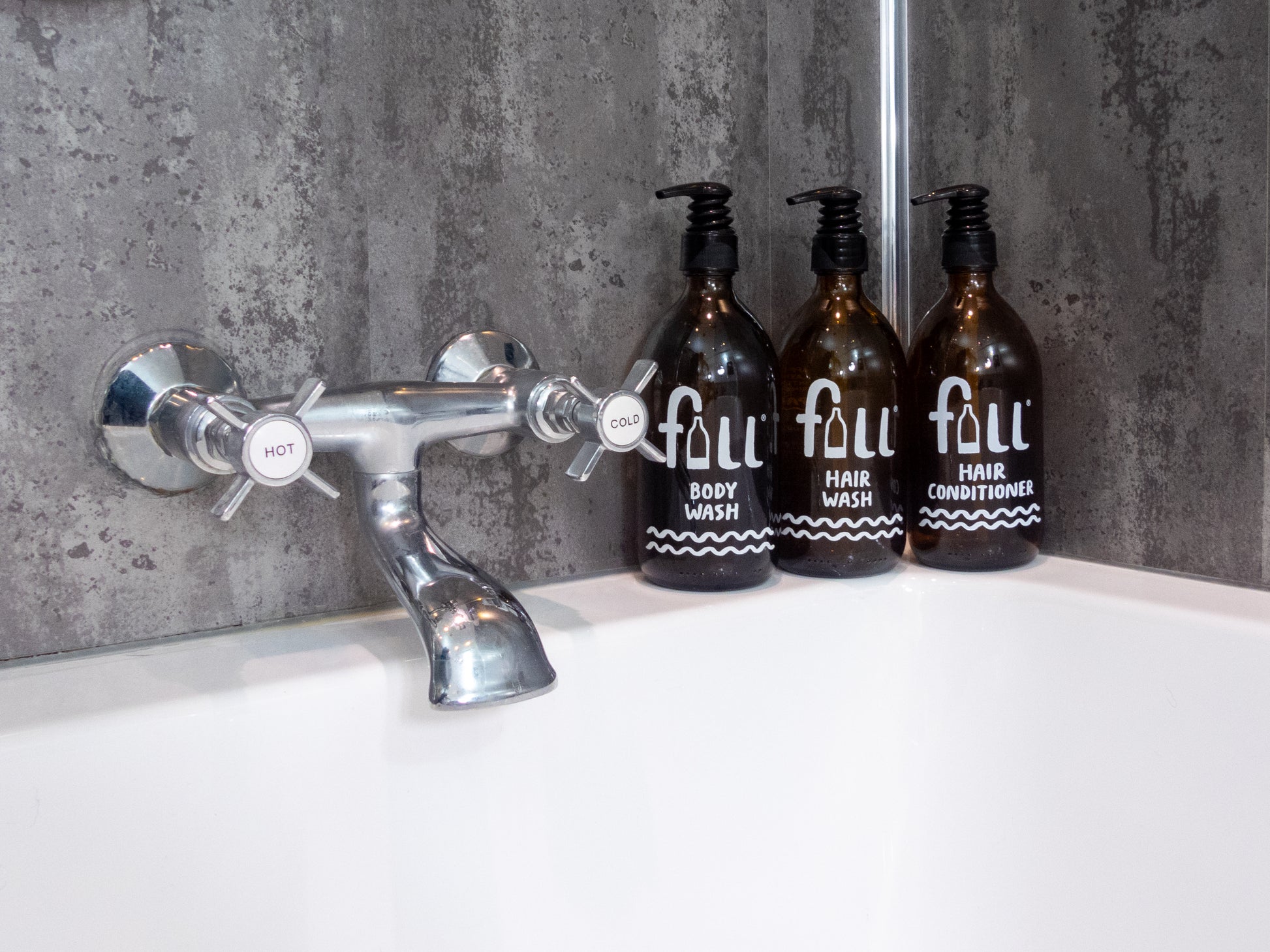 Trio of amber glass bottles on a bath tub containing body wash, shampoo and conditioner. All Vegan and SLS-free