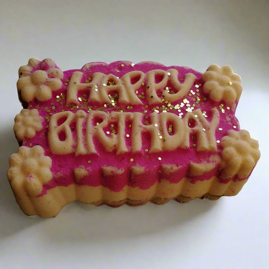 A bath bomb with 'HAPPY BIRTHDAY' text and pink color, scattered with biodegradable glitter.