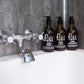 Trio of amber glass bottles on a bath tub containing body wash, shampoo and conditioner. All Vegan and SLS-free